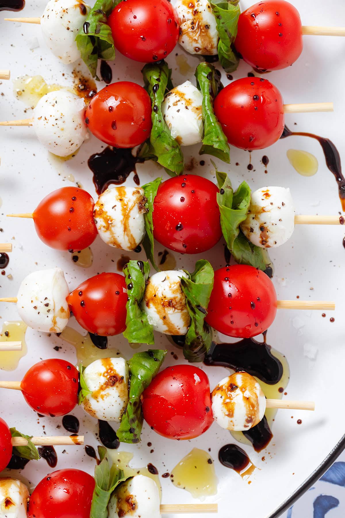 Bamboo skewers with cherry tomatoes, bocconcini, and fresh basil drizzled with olive oil and balsamic glaze, and flaky sea salt on a white plate.