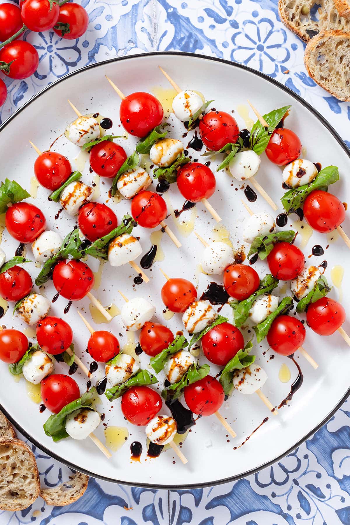 Wooden skewers with cherry tomatoes, bocconcini, and fresh basil drizzled with olive oil and balsamic glaze on a large white platter.