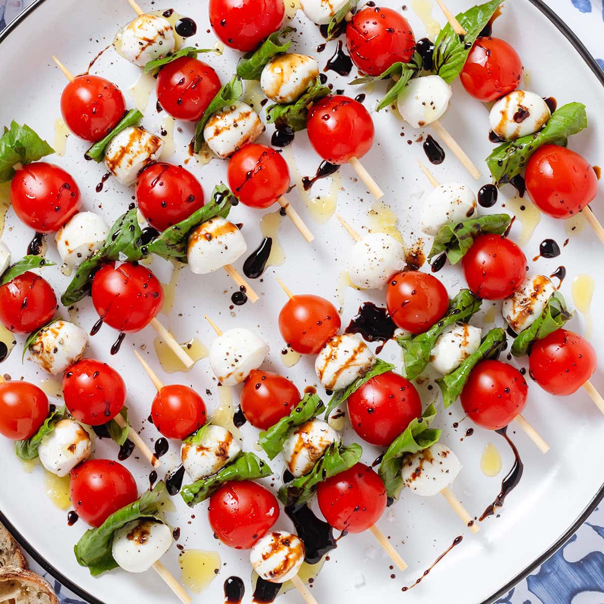 Wooden skewers with cherry tomatoes, bocconcini, and fresh basil drizzled with olive oil and balsamic glaze on a large white platter.