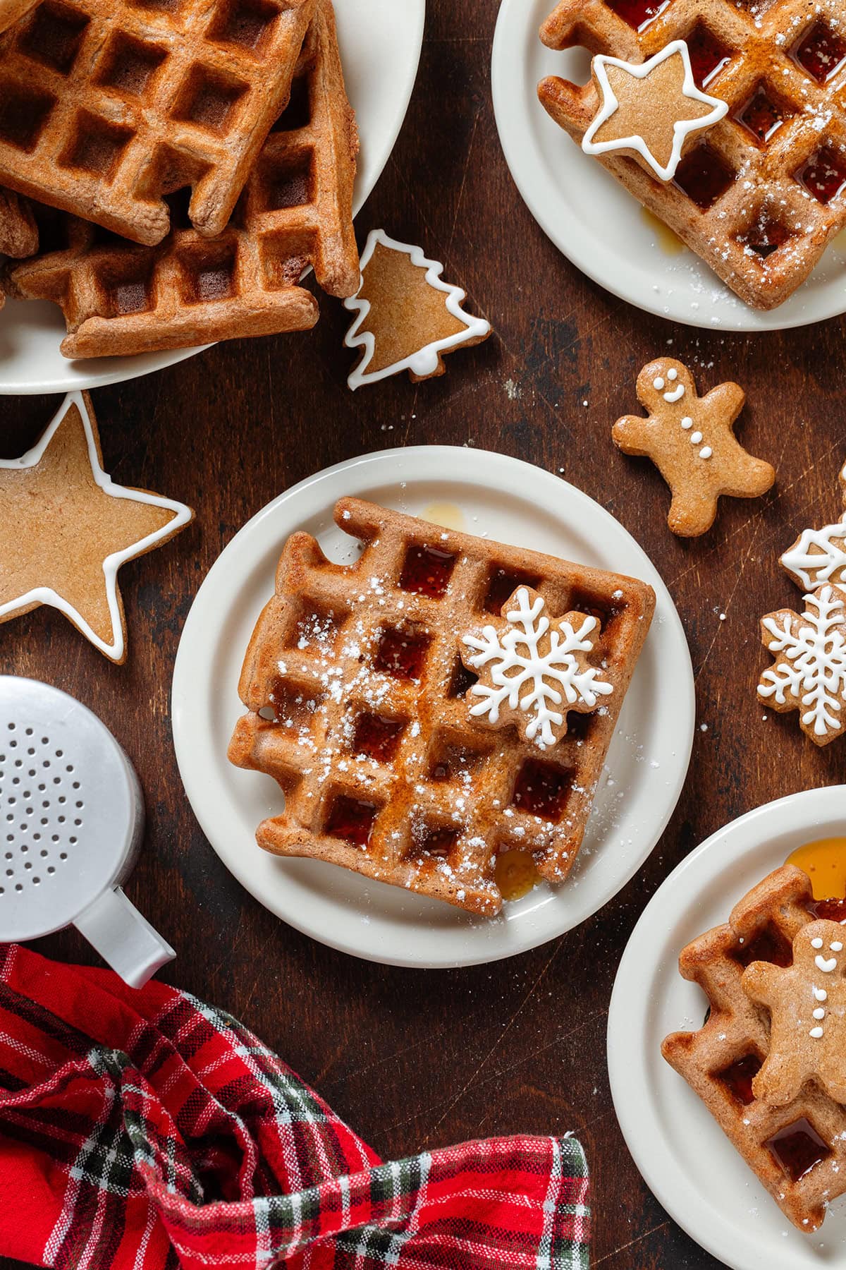 Light brown waffles on small white plates topped with mini gingerbread cookies, a drizzle of maple syrup, and a dusting of powdered sugar.