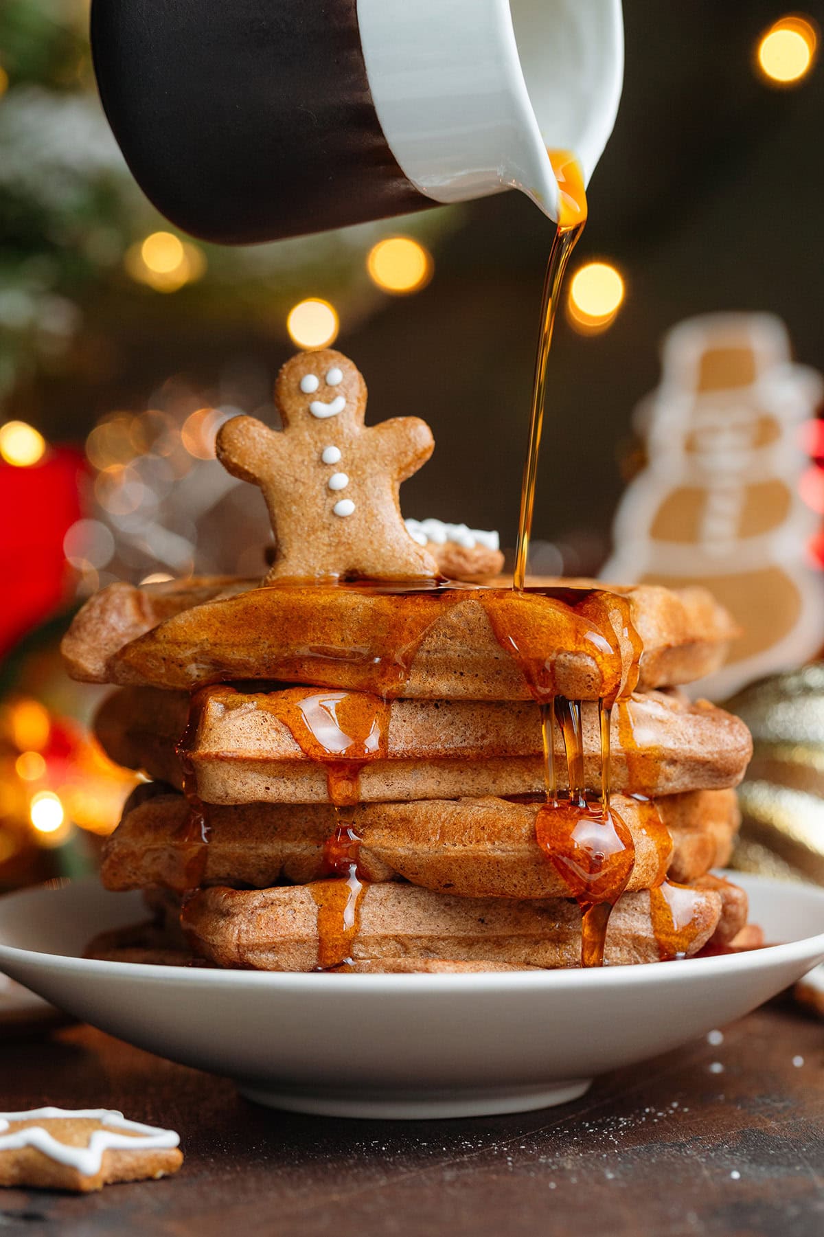 A stack of waffles with a small gingerbread man cookie on top being drizzled with maple syrup.