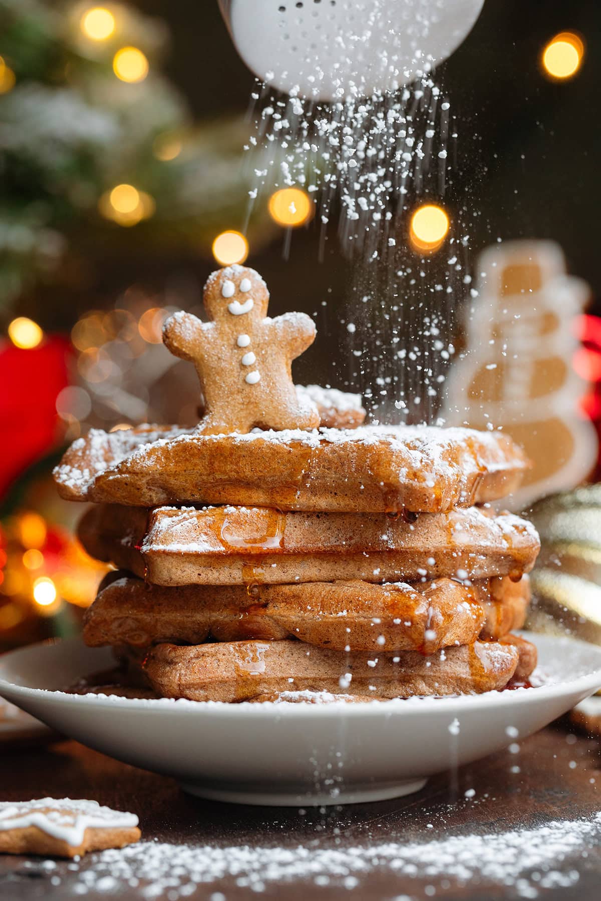 A stack of waffles with a small gingerbread man cookie on top being dusted with powdered sugar.