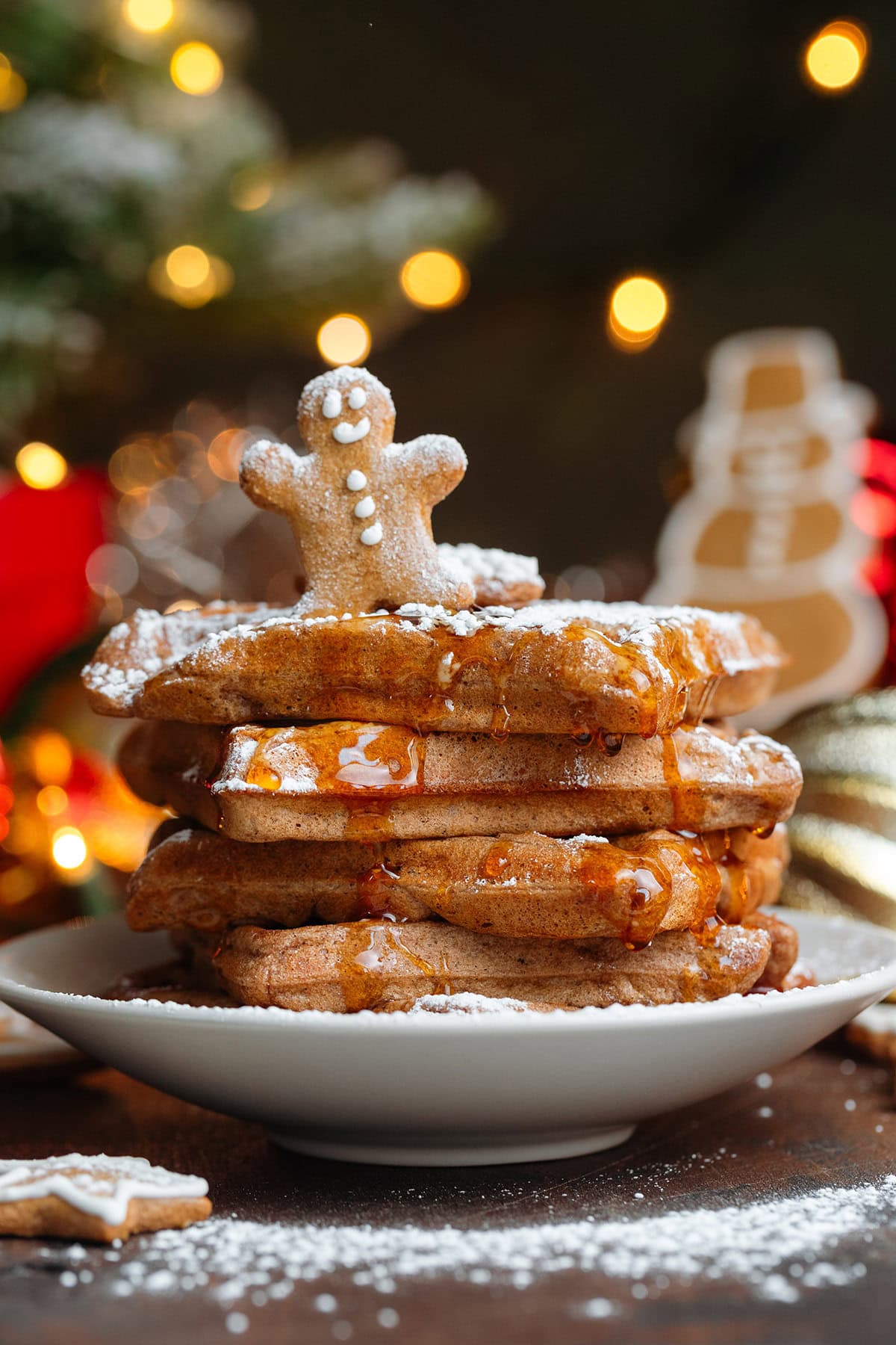 A stack of waffles with a small gingerbread man cookie on top drizzled with maple syrup and dusted with powdered sugar.