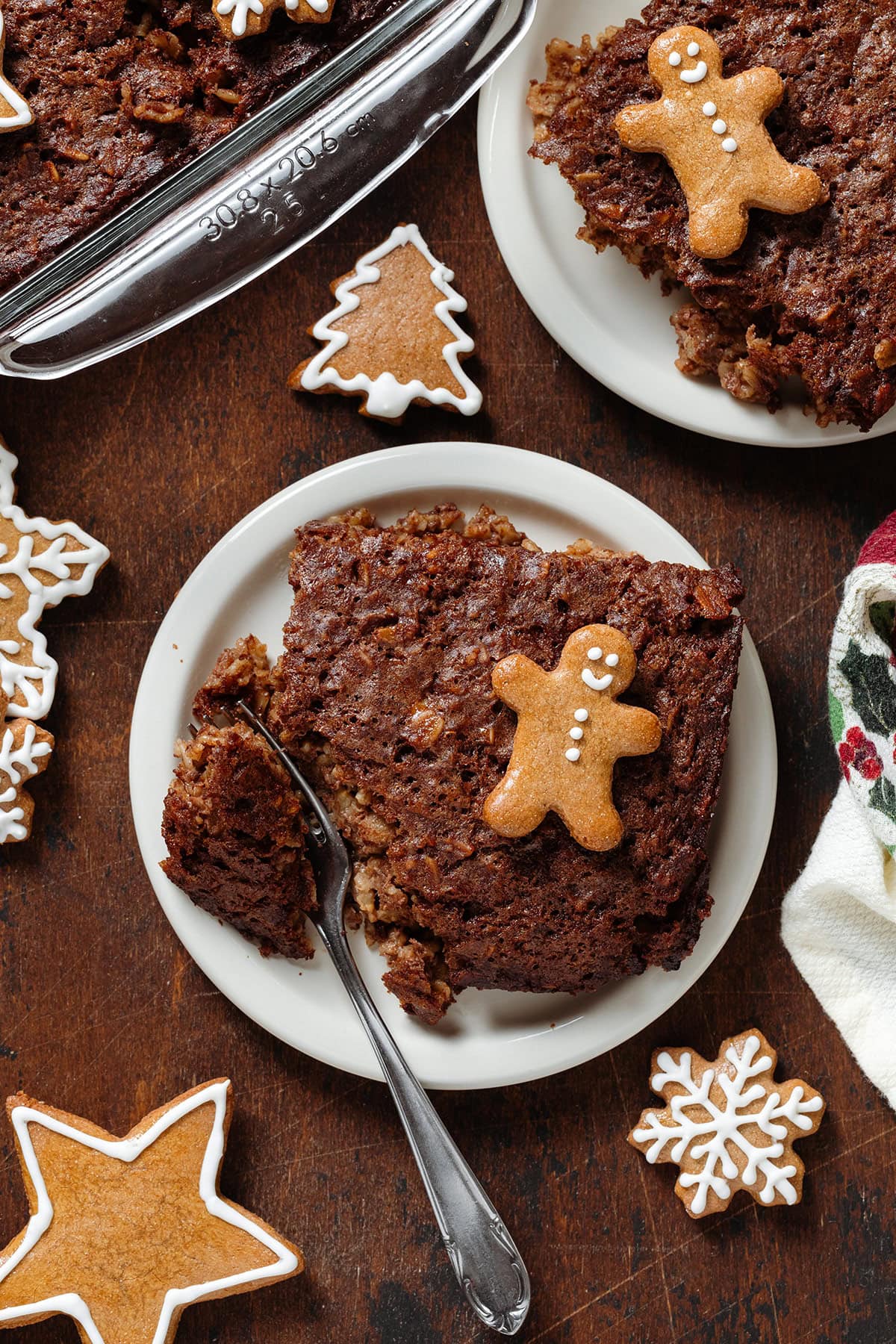 Two squares of baked oatmeal on small white plates garnished with a small gingerbread man cookies and a fork slicing off a piece from one of the plates.