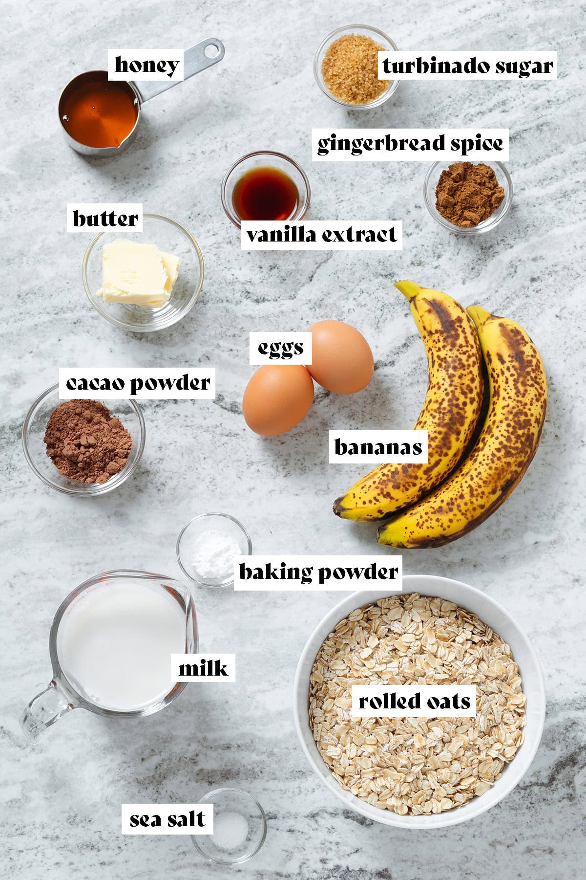 Rolled oats, milk, bananas, eggs, cacao powder, butter, honey, and spices in small glass bowls laid out on a grey background with text overlay.