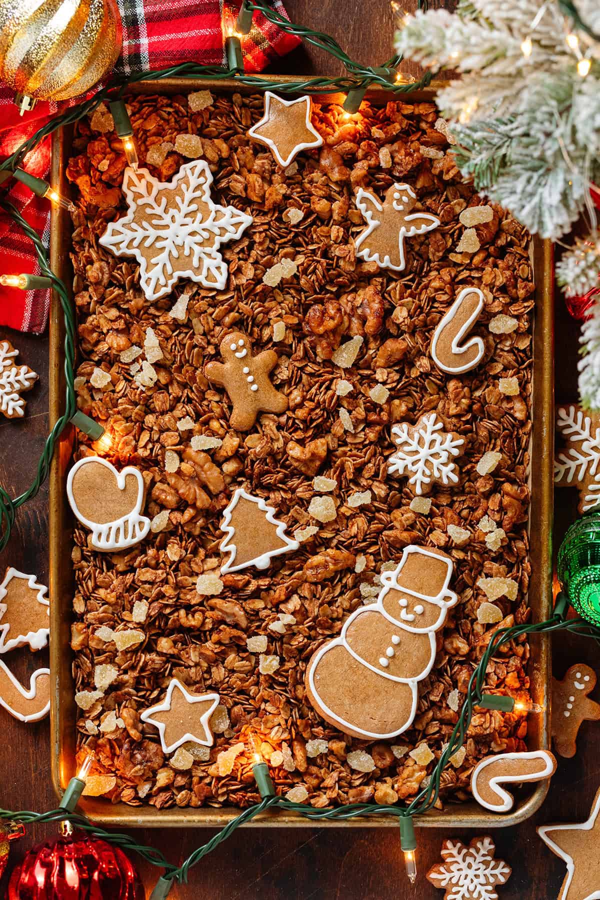 Granola with crystallized ginger on a large baking sheet with gingerbread cookies on the granola surrounded with christmas ornaments and a red tea towel.