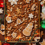 Granola with crystallized ginger on a large baking sheet surrounded with Christmas ornaments and more cookies.