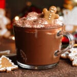 Hot chocolate in a glass mug topped with whipped cream, grated chocolate and a little gingerbread man.