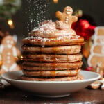 A stack of pancakes with chocolate yogurt and a small gingerbread cookie on top being dusted with powdered sugar.