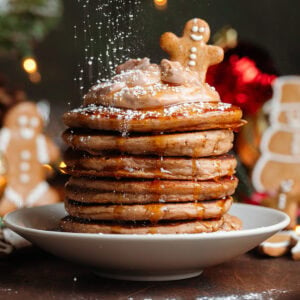 A stack of pancakes with chocolate yogurt and a small gingerbread cookie on top being dusted with powdered sugar.