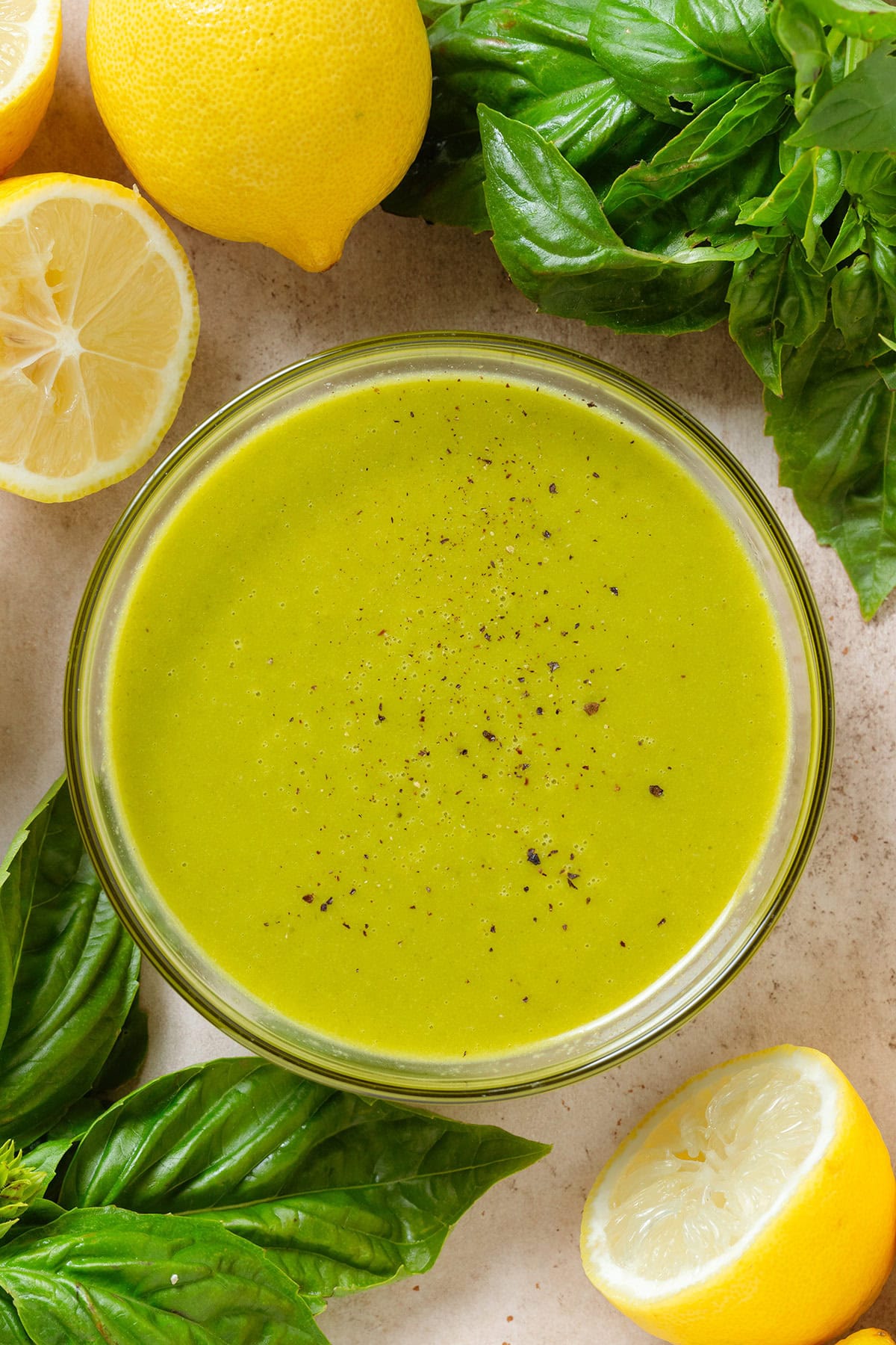 Bright green vinaigrette in a small glass bowl sprinkled with black pepper and with fresh basil and lemons around the bowl.