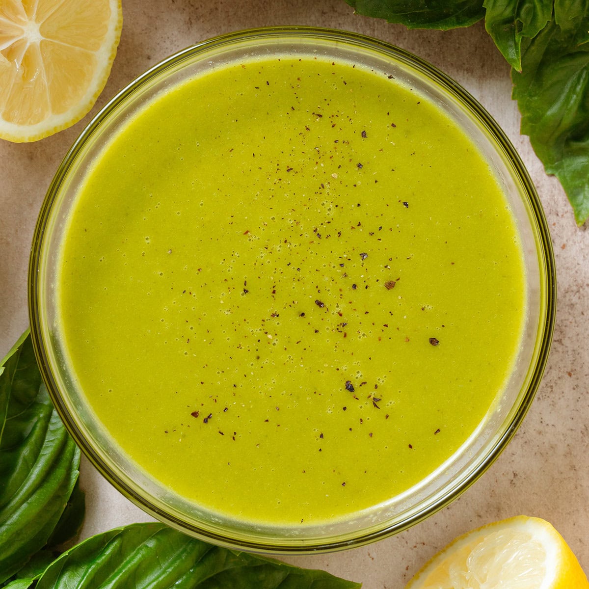 Bright green vinaigrette in a small glass bowl sprinkled with black pepper and with fresh basil and lemons around the bowl.