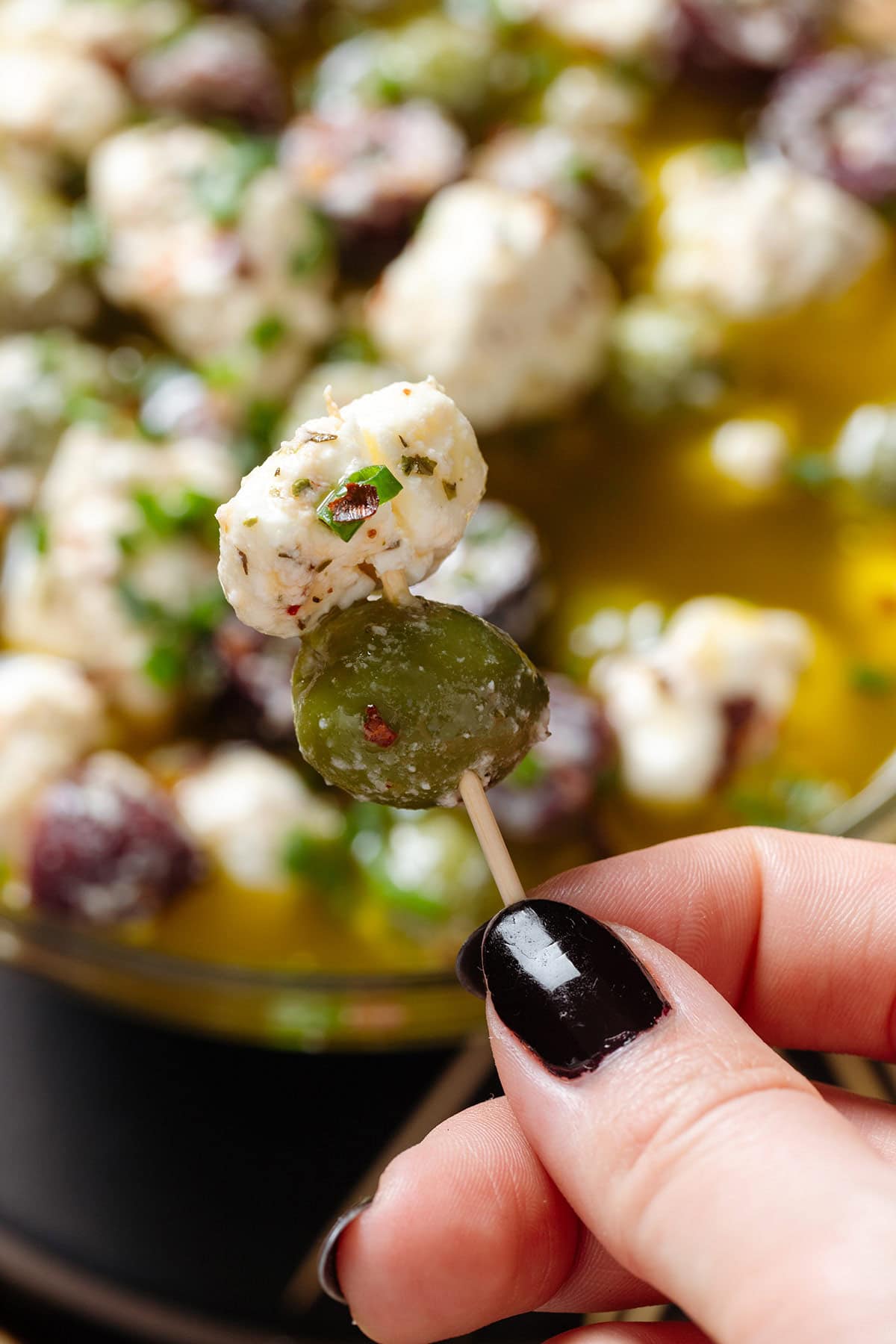 A hand holding a toothpick with a marinated green olive and feta.