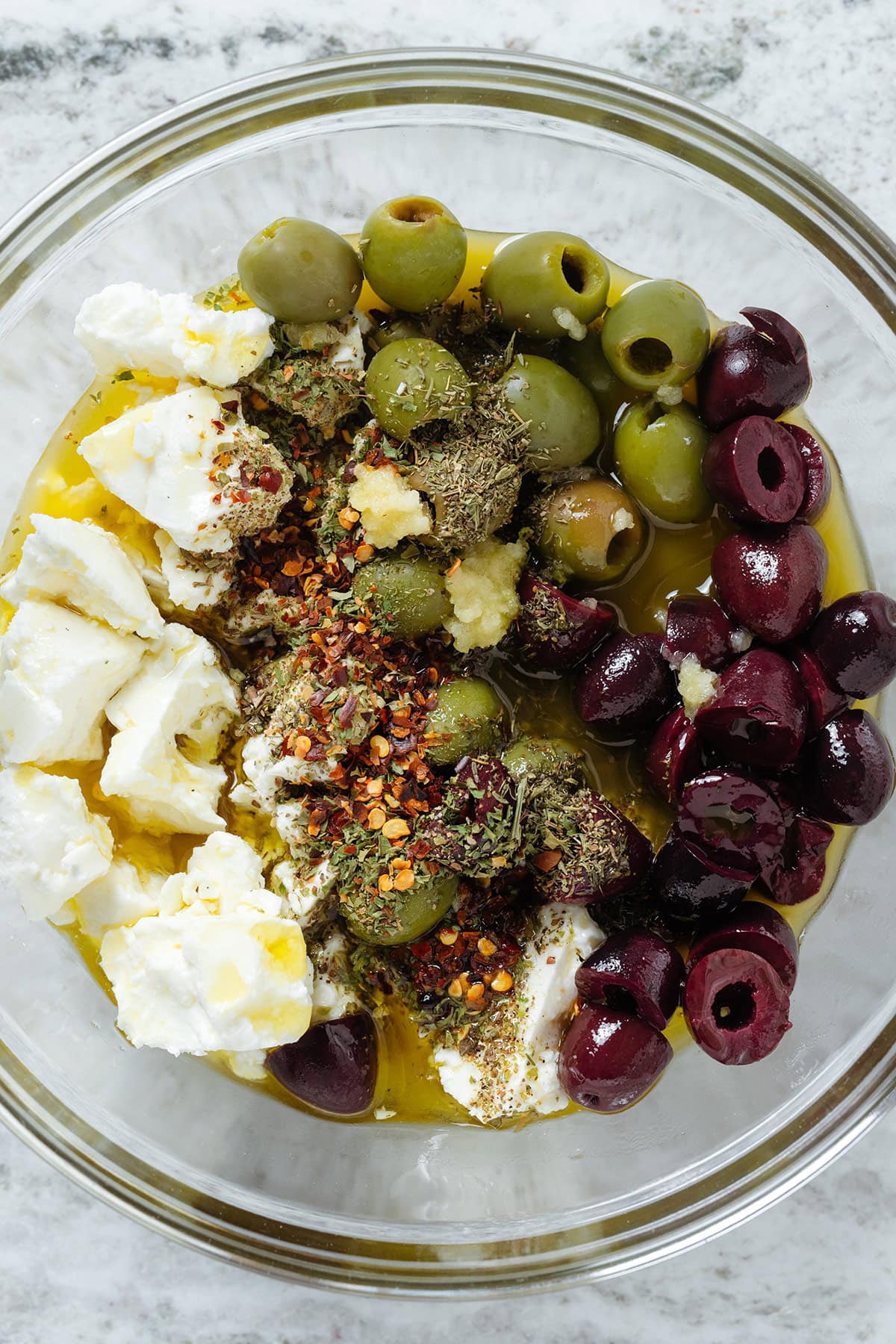 Olives, crumbled feta, spices, minced garlic and olive oil in a glass bowl.