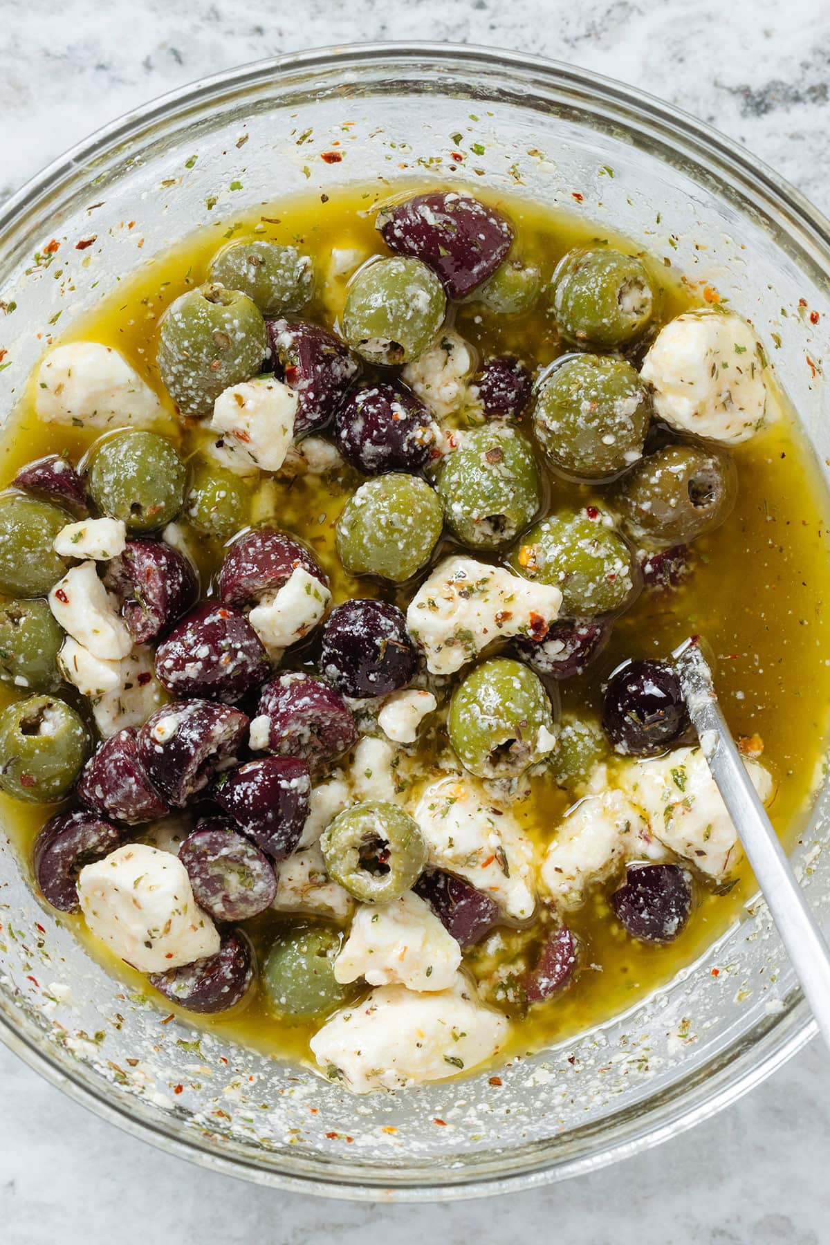 Green and kalamata olives with crumbled feta in a glass bowl marinating with olive oil, spices, and vinegar.