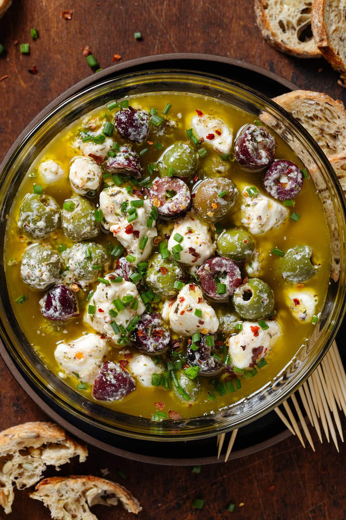 Marinated olives and crumbled feta in olive oil and spices in a glass bowl with toothpicks on the side.