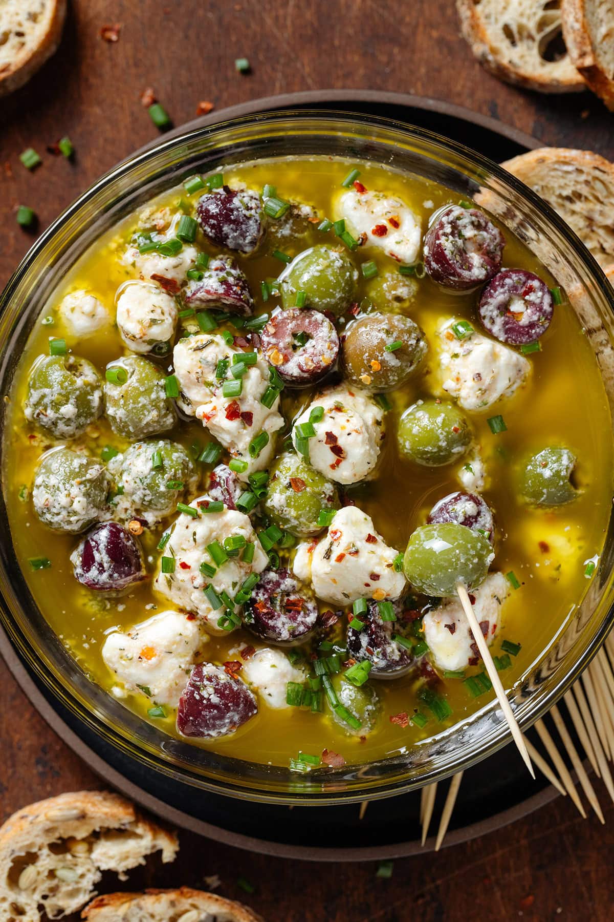 Marinated olives and crumbled feta in olive oil and spices in a glass bowl with toothpicks on the side and one picking out an olive.