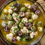 Marinated olives and crumbled feta in olive oil and spices in a glass bowl with toothpicks on the side.