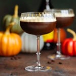 Espresso martini in a pumpkin shaped coupe glass on a dark wooden background with pumpkins behind it.