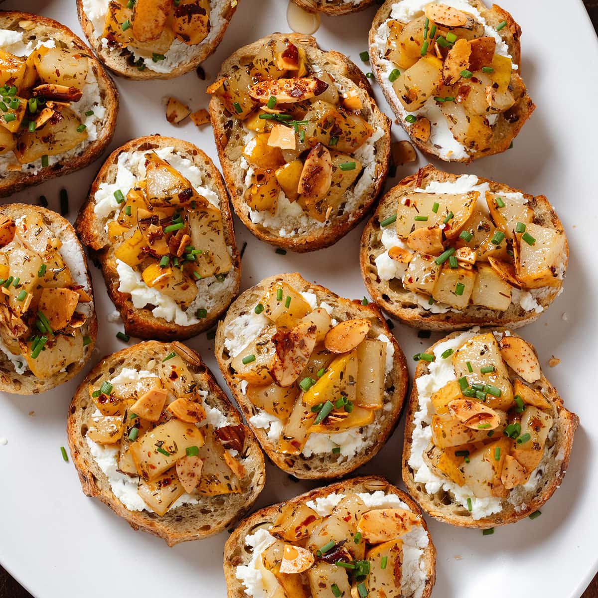 Crostini with soft goat cheese, roasted diced pears, and toasted almonds on a large white serving platter.