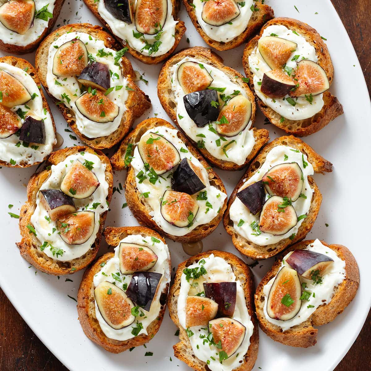 Crostini with whipped ricotta, chopped fresh black figs, and fresh parsley on a large white serving plate.