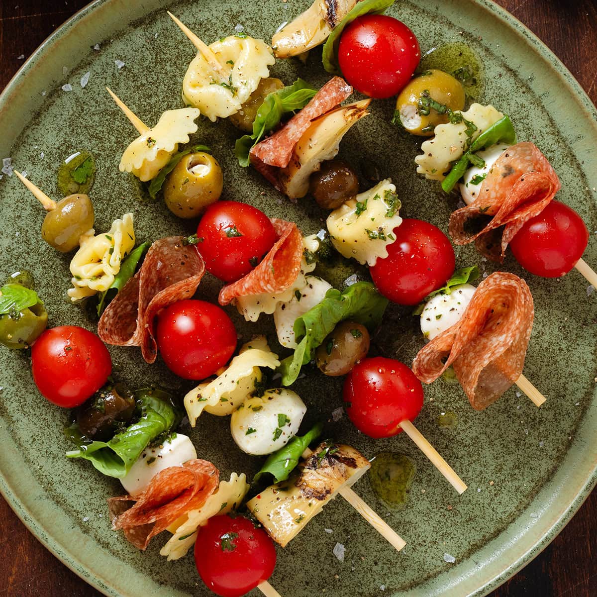 Skewers with cherry tomatoes, mozzarella, tortellini, olives, salami, and other small bites on a green plate.