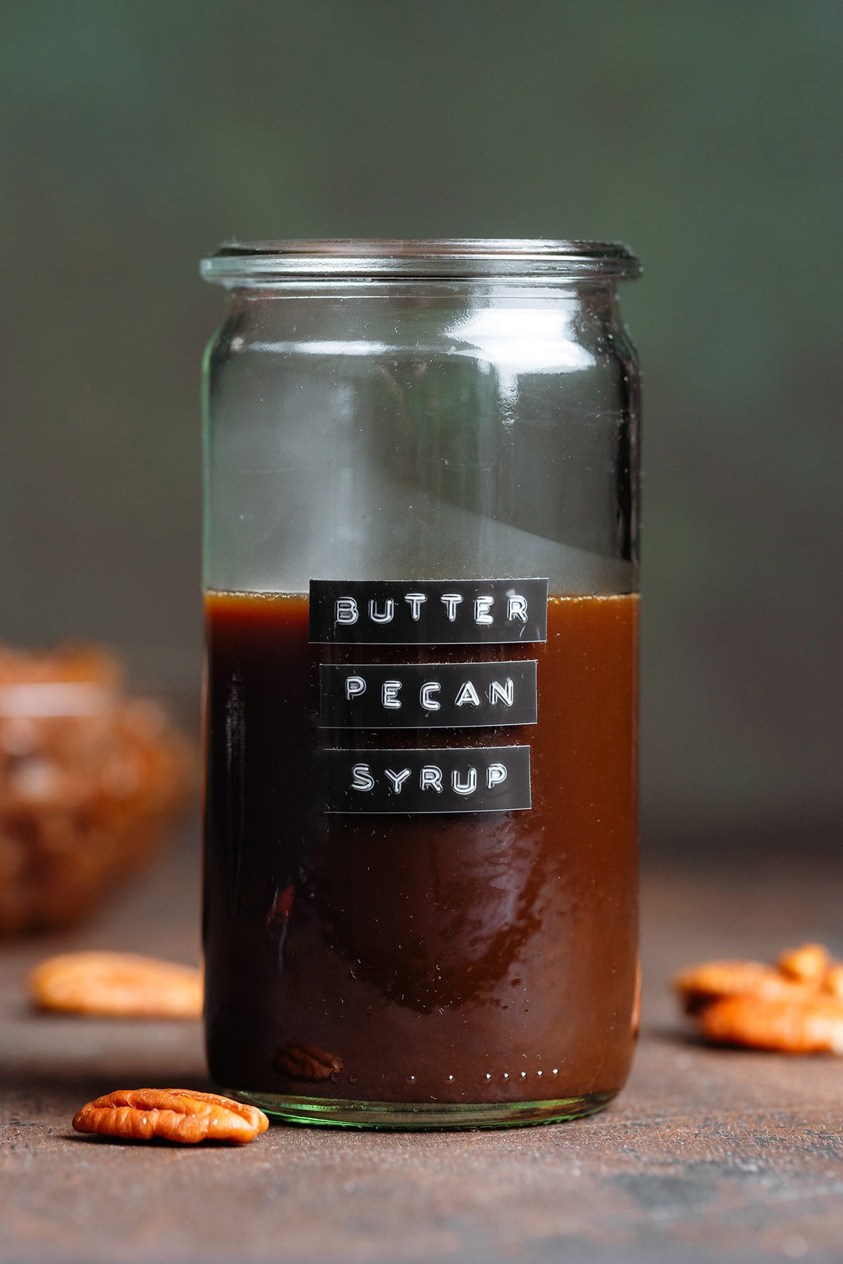 A glass jar with dark brown syrup without a lid with a black embossed label on the side that says butter pecan syrup.