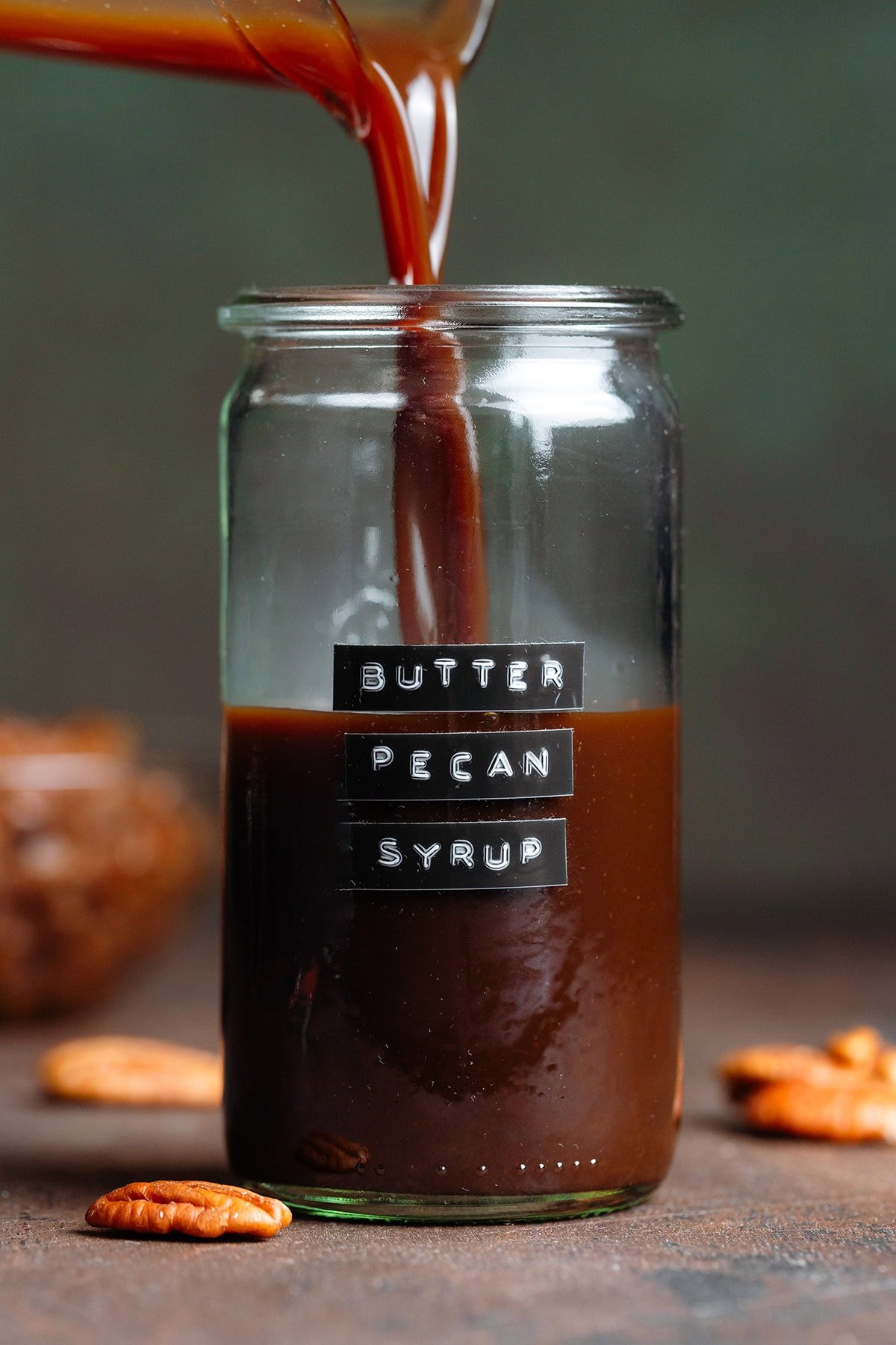 Dark brown syrup being poured into a glass jar with an embossed label that says butter pecan syrup.