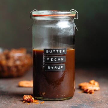 A glass jar with dark brown syrup with a black embossed label that says butter pecan syrup.