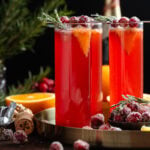 Bright red cranberry mimosa in tall stemless flute glasses garnished with sugared cranberries, fresh rosemary and small orange slices on a gold round platter.