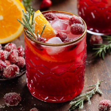 Bright red cranberry drink in a short glass with a gold rim garnished with sugared cranberries, fresh rosemary, and an orange slice.