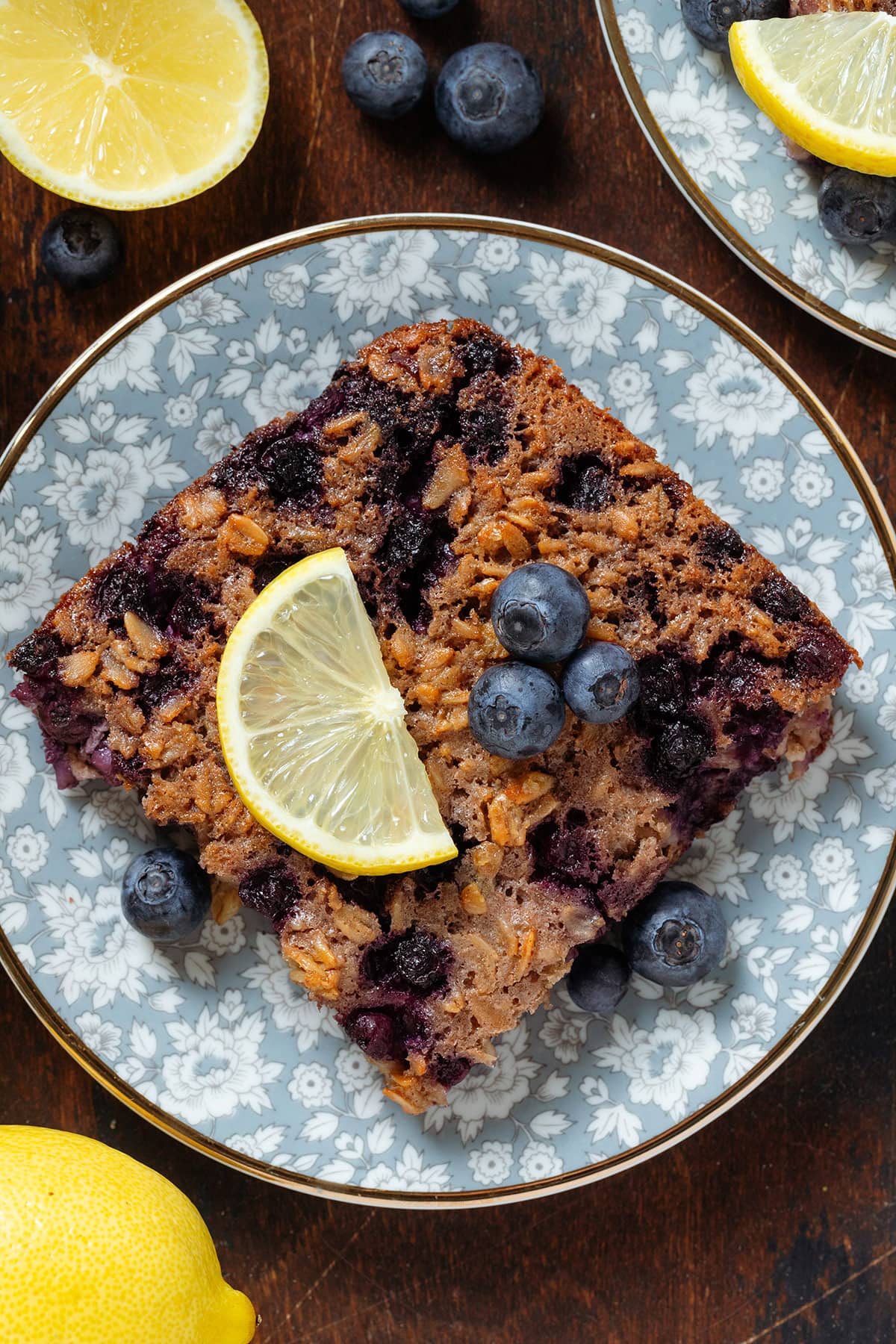 A square of baked oatmeal with blueberries and a lemon slice on top on a blue plate with a gold rim.