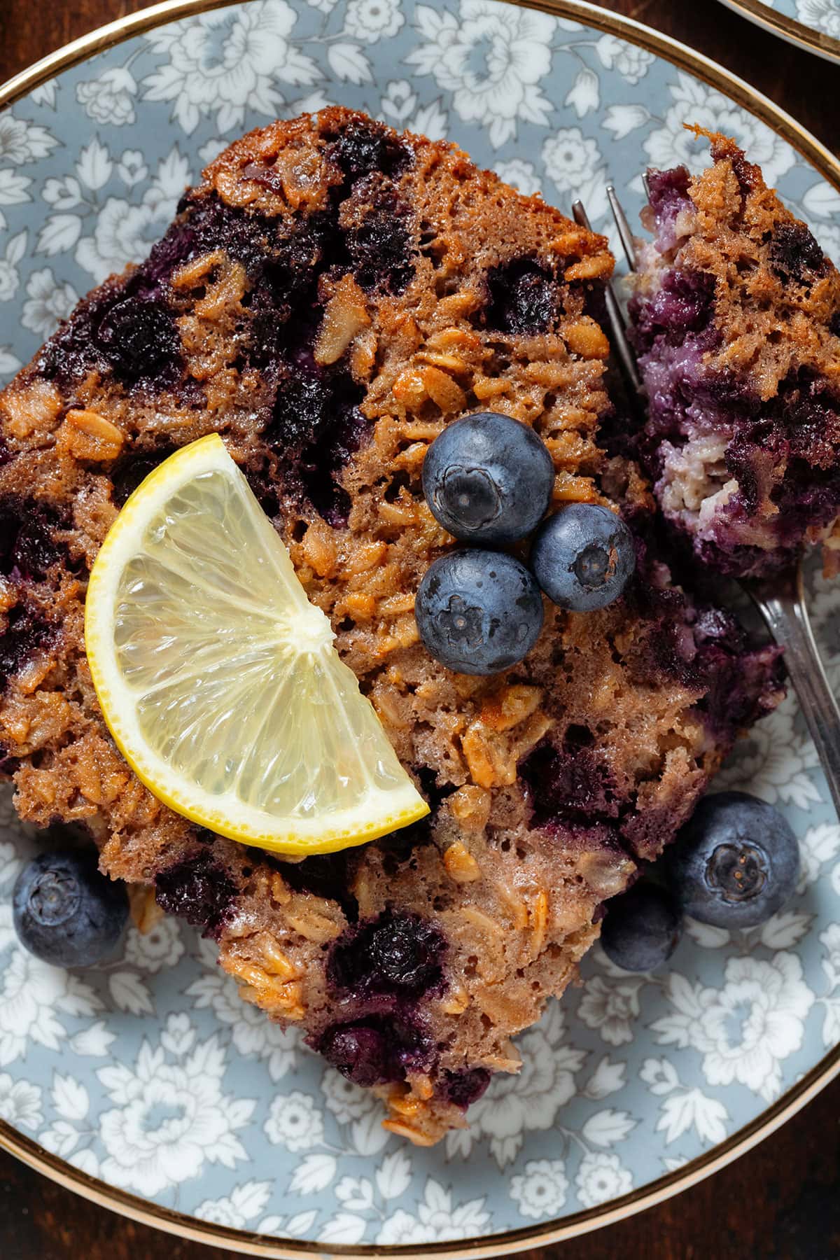 A square of baked oatmeal with blueberries and a lemon slice on top on a blue plate with a fork holding a bite of the oatmeal.