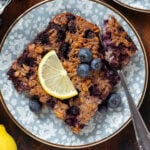 A square of baked oatmeal with blueberries and a lemon slice on top on a blue plate with a fork holding a bite of the oatmeal.