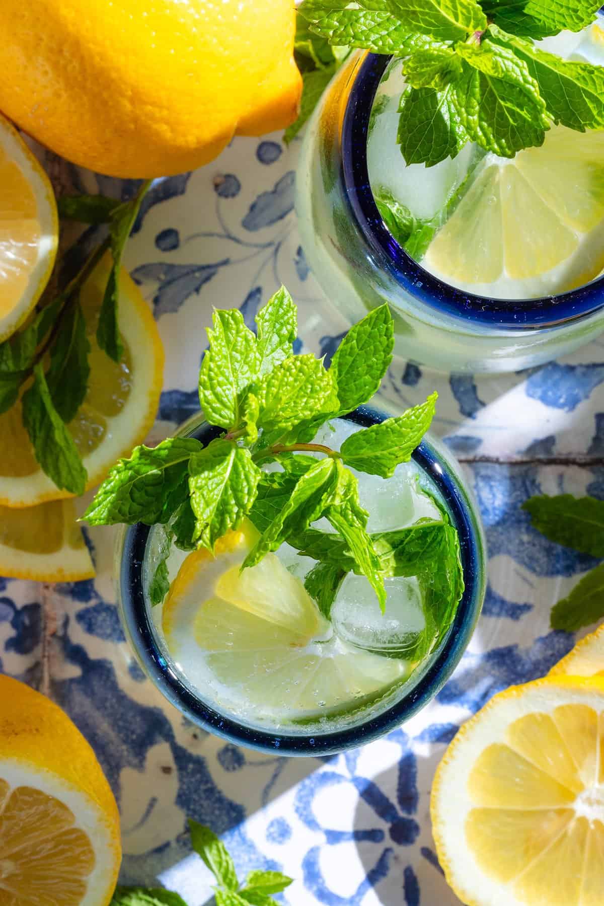 A lemon mocktail in a short glasses with a blue rims garnished with lemon slices and fresh mint on a blue tile background.