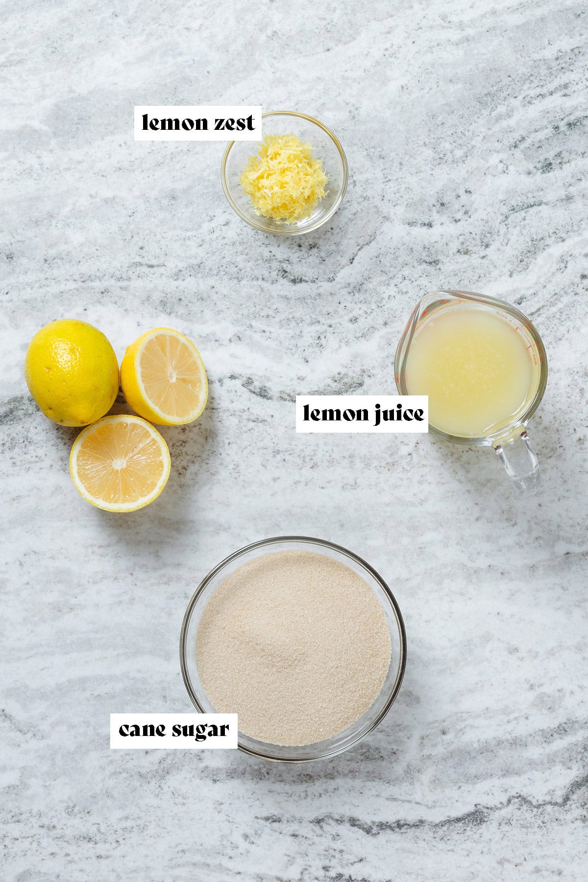 Cane sugar, lemon zest, and lemon juice measured in glass bowls and a measuring cup next to whole lemons on the left.