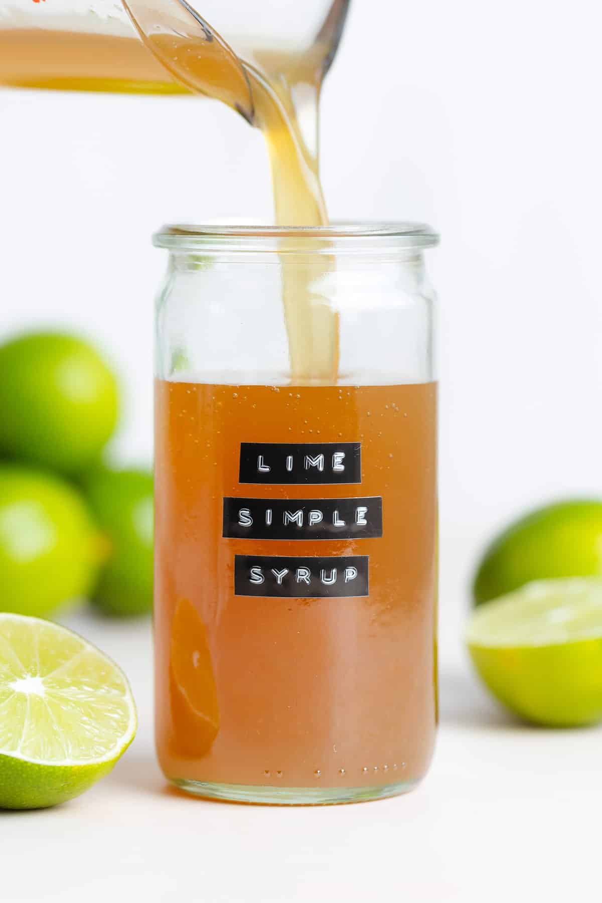 Dark yellow syrup being poured into a tall glass jar with an embossed label that says lime simple syrup.