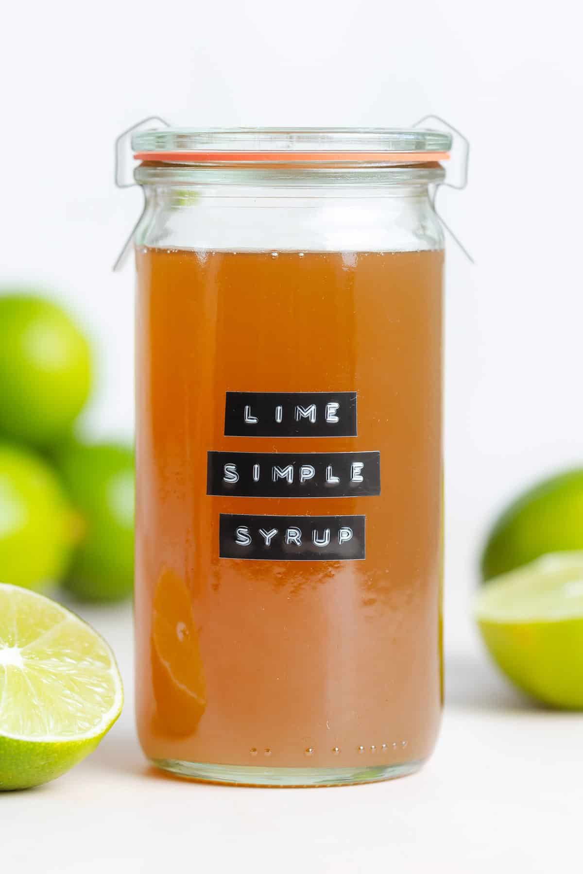 Dark yellow syrup in a glass jar with a black embossed label that says lime simple syrup with limes all around.