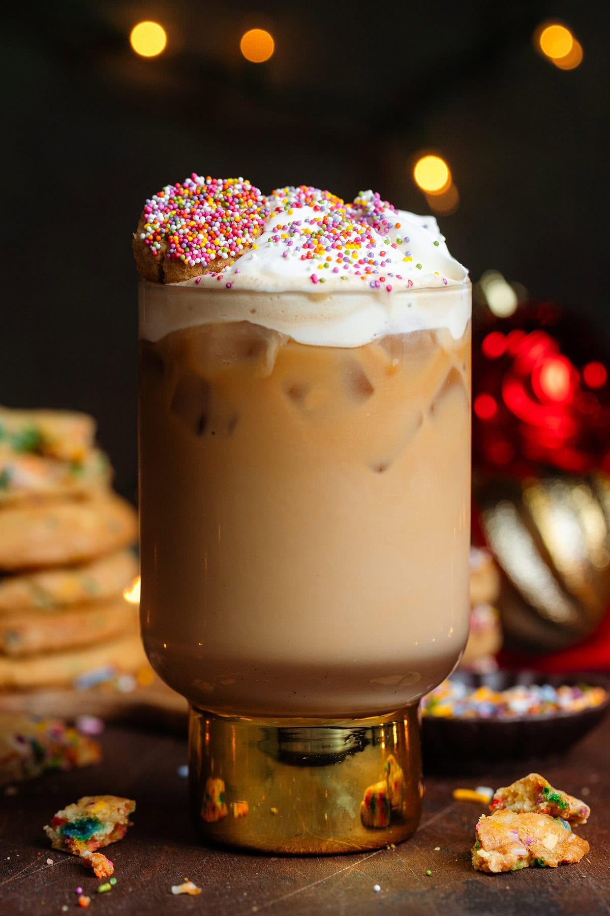 Iced latte in a tall glass with a thick gold stem topped with whipped cream, sprinkles, and a small cookie.