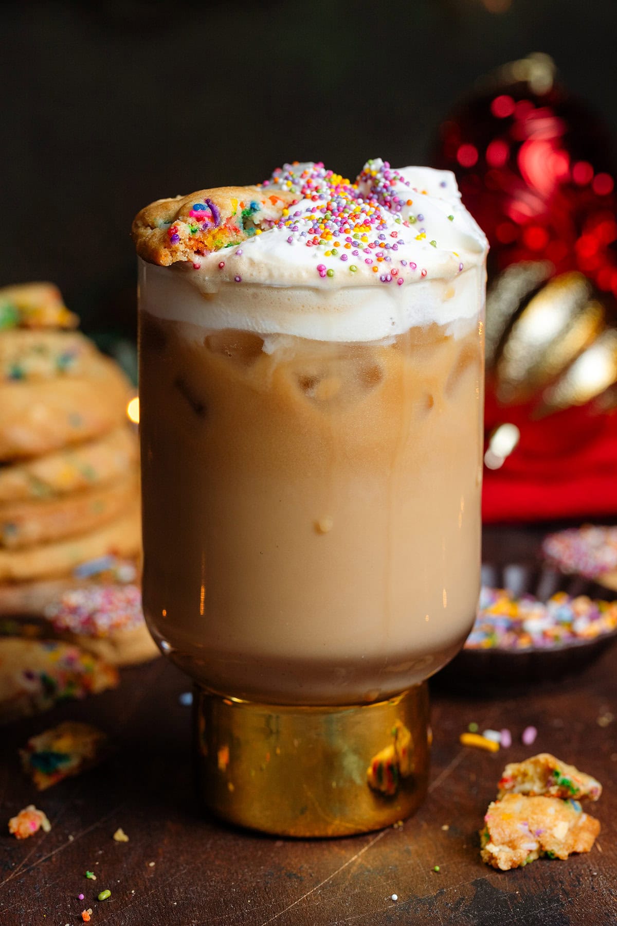 Iced latte in a tall glass with a thick gold stem topped with whipped cream, sprinkles, and a quarter of a funfetti cookie.