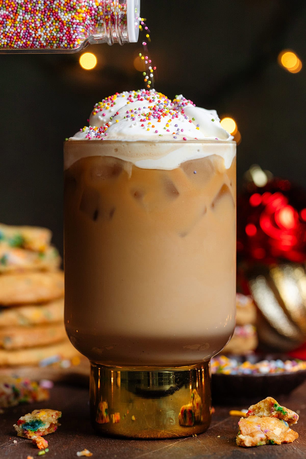 An iced latte with whipped cream being sprinkled with colorful mini sprinkles.