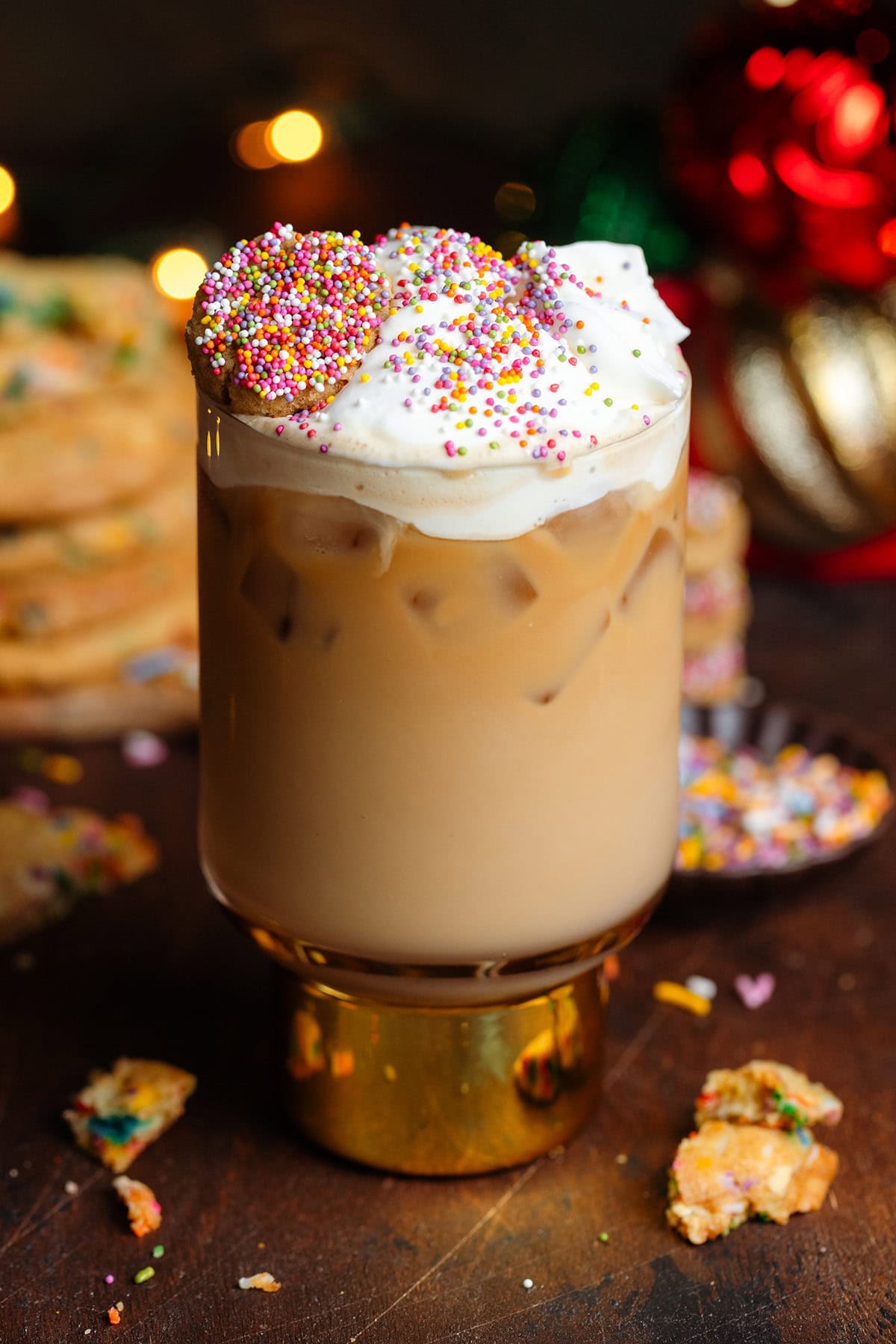 Iced latte in a tall glass with a thick gold stem topped with whipped cream, sprinkles, and a small cookie on top.