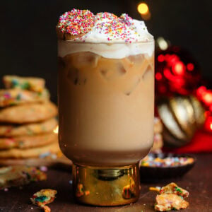 Iced latte in a tall glass with a thick gold stem topped with whipped cream, sprinkles, and a small cookie.