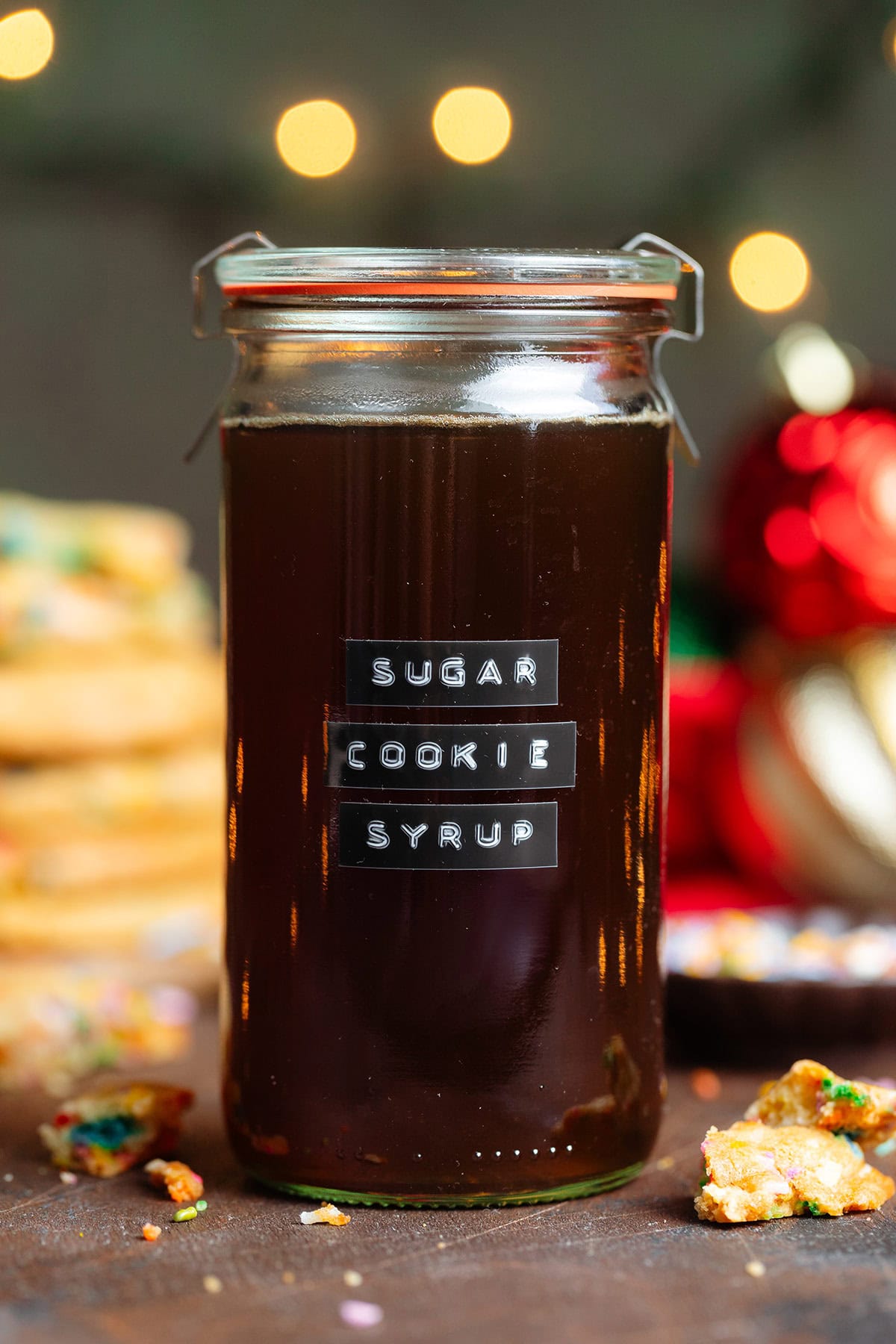 Dark brown syrup in a glass jar with a lid and an embossed label that says sugar cookie syrup with cookies in the background.