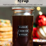 Dark brown syrup in a glass jar with a lid and an embossed label that says sugar cookie syrup with cookies in the background.