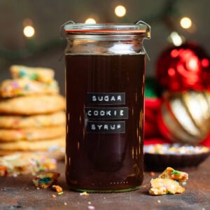 Dark brown syrup in a glass jar with a lid and an embossed label that says sugar cookie syrup with cookies in the background.