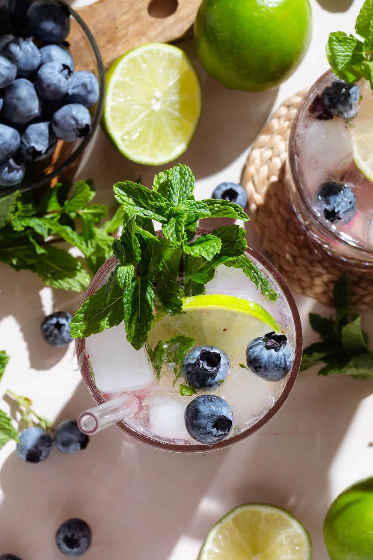 A cocktail in a tall glass garnished with blueberries, fresh mint, and lime slices shot from above.