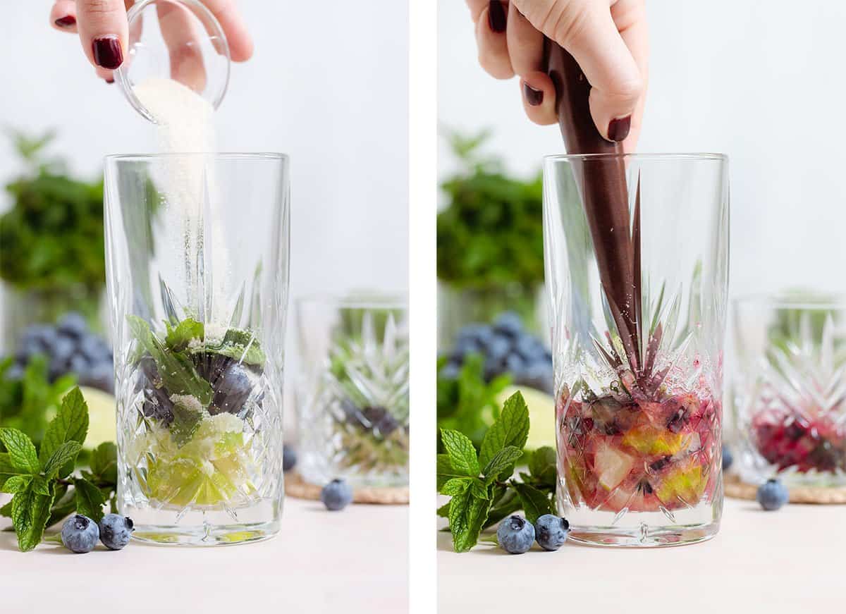 Cane sugar being added to a tall glass with blueberries, mint, and chopped lime and muddled with a muddler.