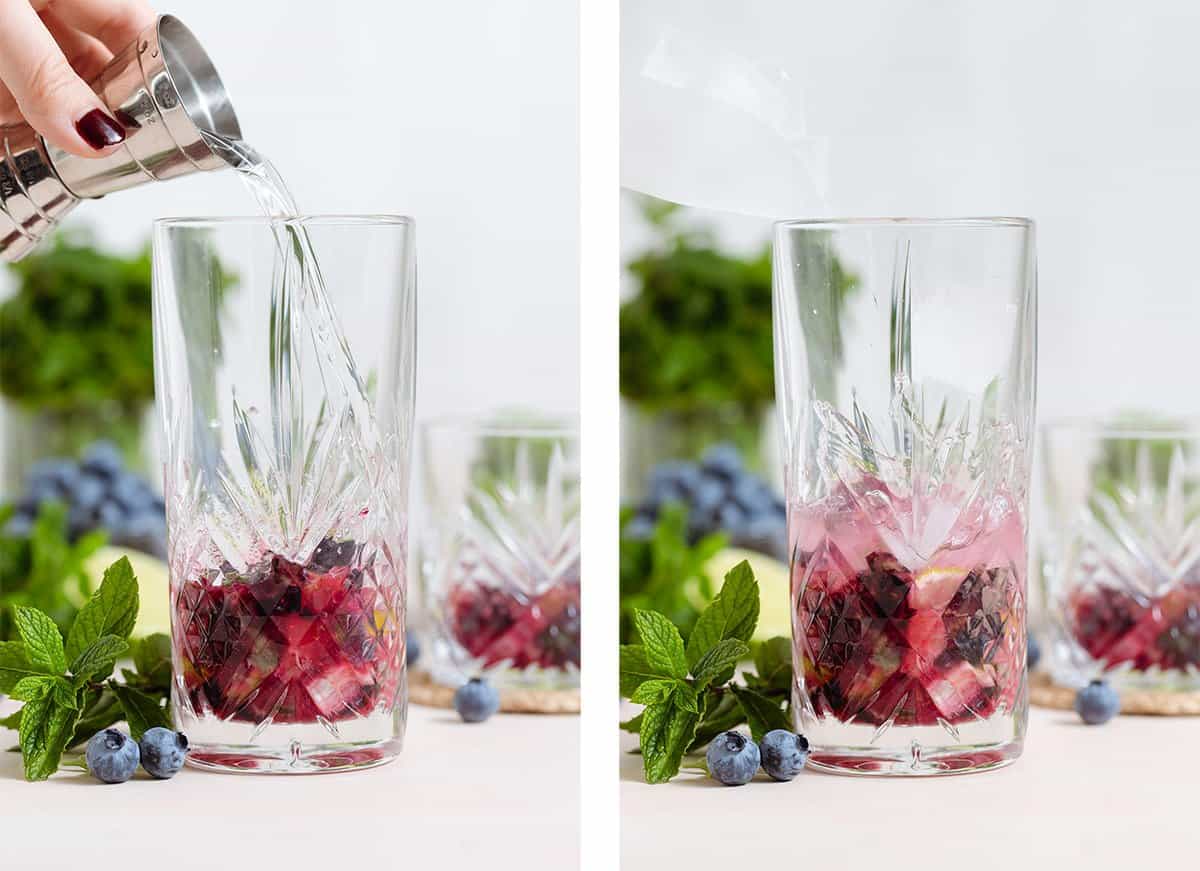 White rum being poured into a tall glass with mashed blueberries and mint and ice being added.