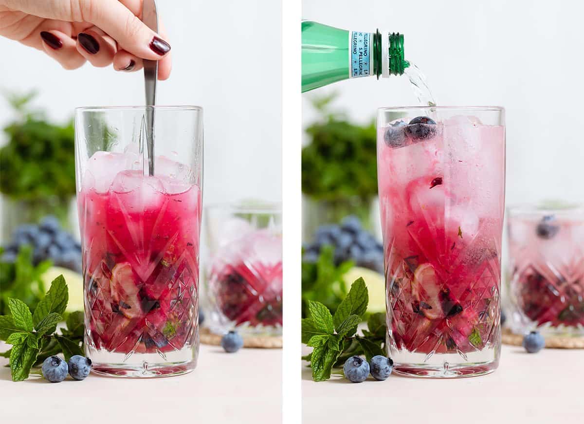 A bright purple cocktail in a tall glass being stirred with a long spoon and topped with sparkling water.